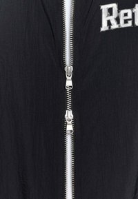 Schwarze Reißverschlussjacke aus strukturiertem Stoff, mit doppelten silbernen Reißverschlüssen und einem aufgedruckten Logo im oberen Bereich. Sauberes, minimalistisches Design.