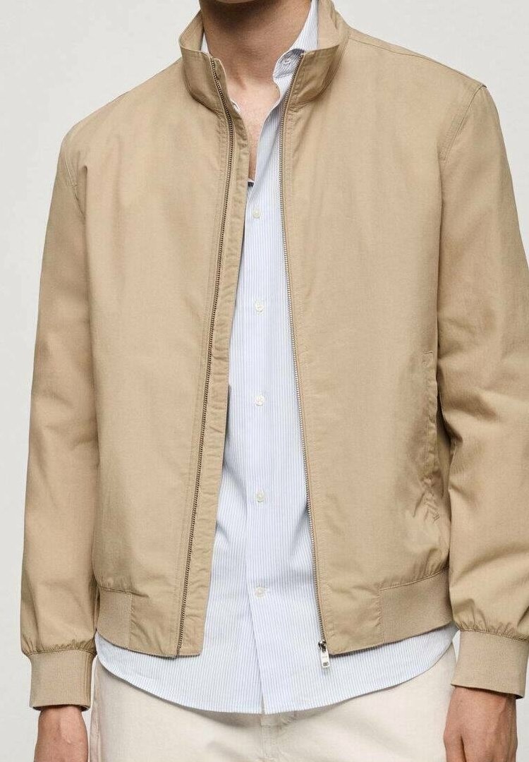 Homme portant une veste beige zippée par-dessus une chemise à rayures bleu clair boutonnée et un pantalon clair, debout devant un fond uni.