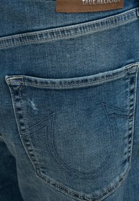 Niebieskie dżinsy z tylną kieszenią ozdobioną charakterystycznym haftem w kształcie podkowy oraz skórzaną naszywką z marką "True Religion."