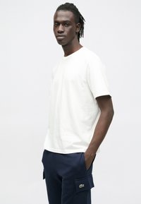 Biele bavlnené tričko s krátkymi rukávmi a okrúhlym výstrihom, nosené s námorníckymi nákladnými nohavicami s logom zelenej značky na vrecku.