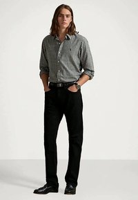 Camisa de botões cinza e branca às quadrículas, calças pretas e sapatos pretos. Relógio no pulso, corte reto, mangas compridas enroladas até aos cotovelos.