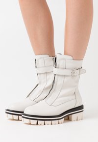Botas de tobillo blancas de cuero suave con cierre de cremallera, acentos de correas anchas y suelas gruesas con rayas en negro y crema.