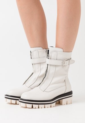 Bottes blanches à la cheville en cuir lisse avec fermeture éclair, accents de larges sangles et semelles épaisses avec des rayures noires et crème.
