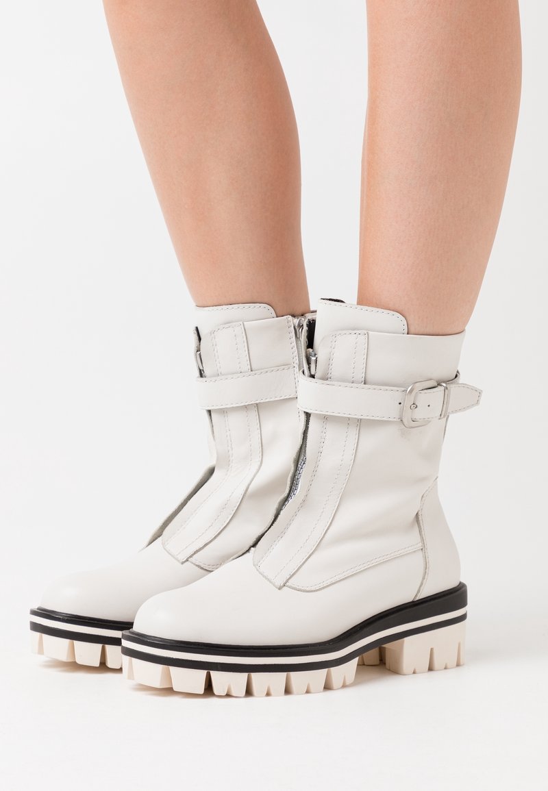 Botas de tobillo blancas de cuero suave con cierre de cremallera, acentos de correas anchas y suelas gruesas con rayas en negro y crema.
