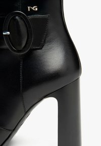 Stivale alto in pelle nera con tacco, caratterizzato da un dettaglio a fibbia tondeggiante, texture liscia e silhouette elegante con finitura lucida.