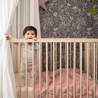 Træcradle med lodrette spær, lys beige finish. Blød pink sengetøj nedenunder. Barn iført en neutralfarvet dragt, lænet over kanten af krybben.