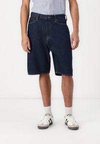 Shorts din denim de culoare bleumarin închis, cu o croială confortabilă, având închidere cu nasture, cinci buzunare și detalii de cusătură subtile. Purtați cu adidași albi.
