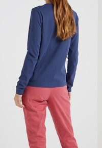 Hedvábný bavlněný top s dlouhým rukávem v námořnické modři, spárovaný s růžovými kalhotami, s kulatým výstřihem a přiléhavými rukávy, zobrazený zezadu.