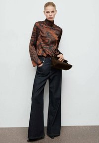 Top de cuello alto con estampado en bronce combinado con pantalones vaqueros anchos en denim oscuro. La modelo sostiene un bolso de mano marrón oscuro. Tela suave y silueta estructurada.