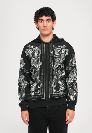 Black zip-up hoodie featuring ornate silver floral patterns and text accents. Material appears smooth, with ribbed cuffs and hem.