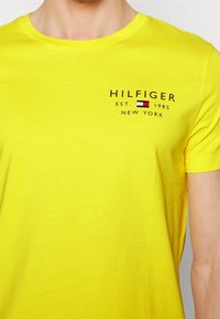 Osoba oblečená v jasně žlutém tričku s nápisem "HILFIGER EST. 1985 NEW YORK" a malou červenou, bílou a modrou vlaječkou na hrudi.