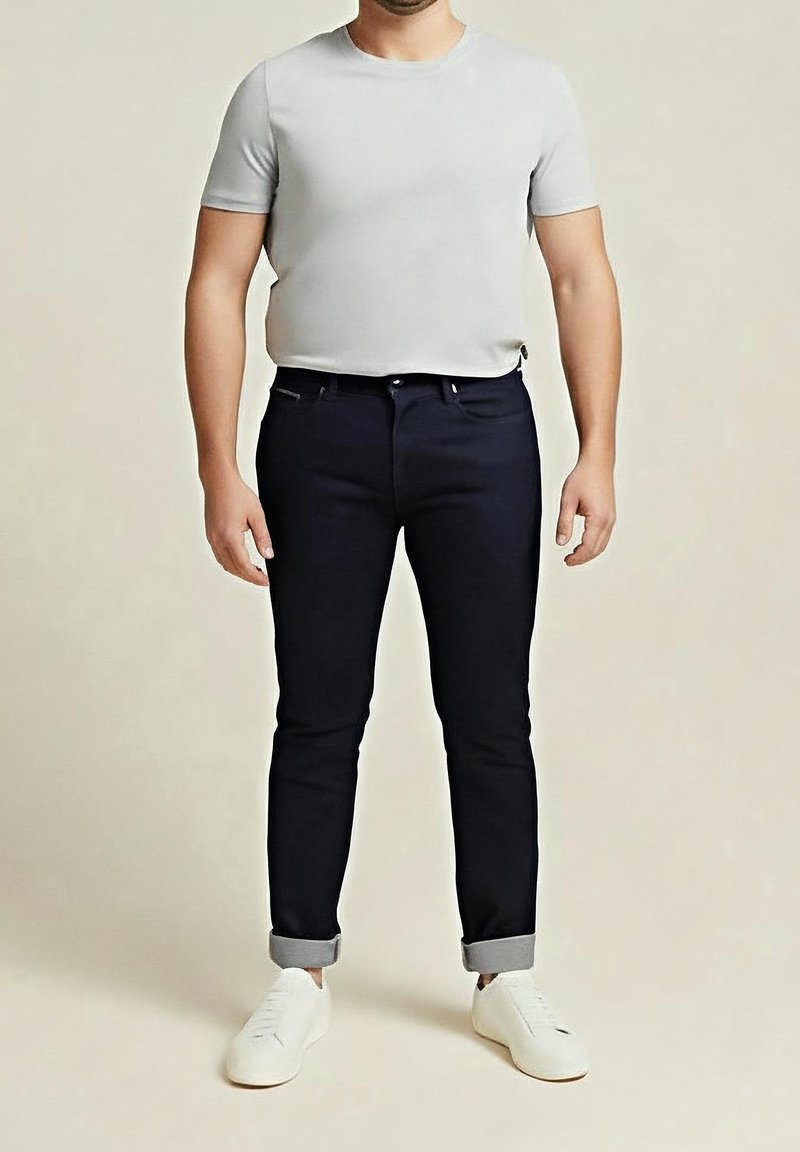 Homme portant un t-shirt gris clair à manches courtes, un jean foncé retroussé et des baskets blanches, debout devant un fond neutre.