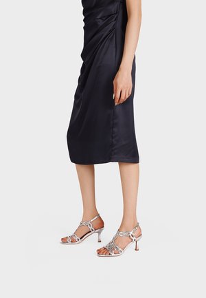 Donna che indossa una gonna midi in satin nera e sandali con tacco argento con dettagli di anelli circolari, in piedi con una mano rilassata lungo il corpo.