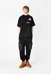 Camiseta negra de gran tamaño con detalle de bolsillo y diseño floral, combinada con pantalones cargo negros con cremalleras y botas naranjas.