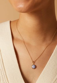 Collier en or avec un pendentif en pierre lavande ronde. La chaîne est fine, avec une texture délicate, reposant sur l'encolure d'un pull de couleur claire.