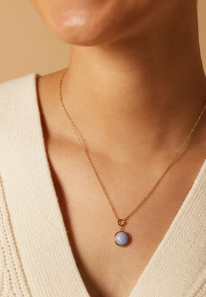 Collier en or avec un pendentif en pierre lavande ronde. La chaîne est fine, avec une texture délicate, reposant sur l'encolure d'un pull de couleur claire.