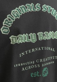 Sort t-shirt med grøn grafisk tekst, der har fed skrift og indviklede mønstre. Inkluderer "est." med et dekorativt emblem nedenfor.