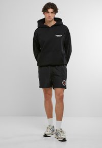 Černá mikina s kapucí a logem, doplněná o černé šortky s červenou grafikou. Model má na sobě bílé kotníkové ponožky a tenisky.