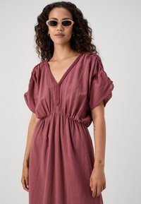 Robe ample à décolleté en V profond dans une teinte rose tamisé, fabriquée en tissu léger. Présente une taille élastique froncée et des manches courtes bouffantes, avec un détail texturé.