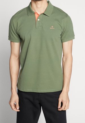 Man draagt een olijfgroene polo met korte mouwen en een klein logo op de linkerborst, gecombineerd met een zwarte broek, staand tegen een effen achtergrond.