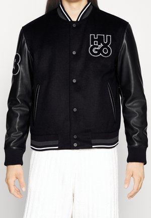 Schwarze Collegejacke mit Lederärmeln, Druckknöpfen, "HUGO"-Logo auf der Brust, Aufnäher am linken Arm, getragen über weißen, strukturierten Hosen.