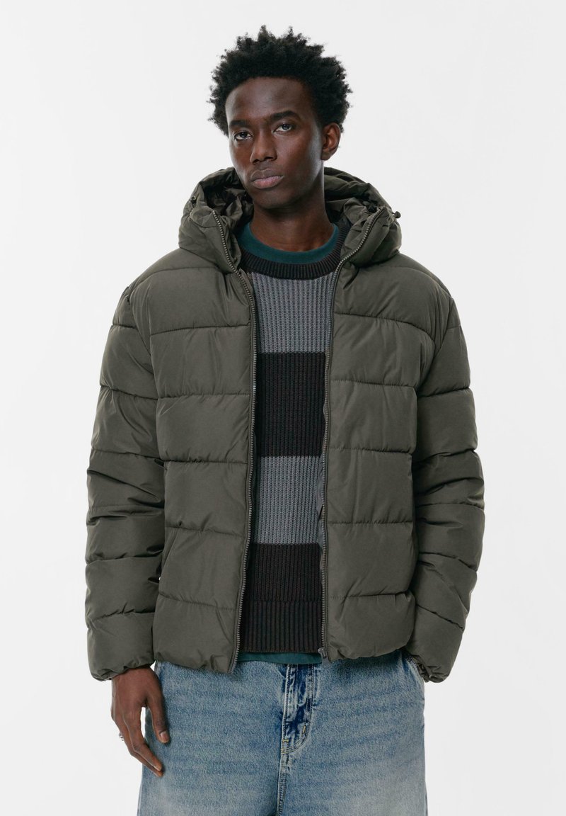 Doudoune à capuche vert olive, avec texture matelassée, fermeture éclair frontale et coupe décontractée. Portée sur un pull rayé et un jean.