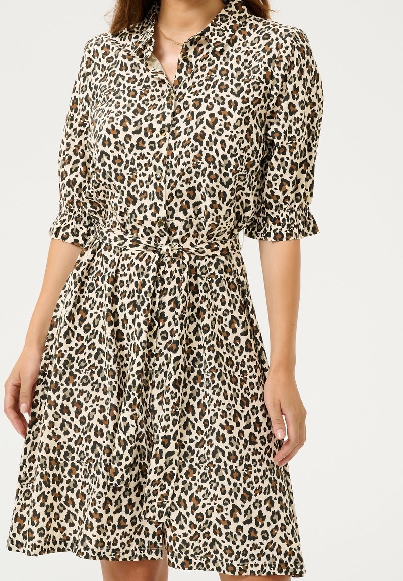 Femme portant une robe boutonnée longueur genoux, imprimé léopard, manches courtes bouffantes et ceinture nouée à la taille.