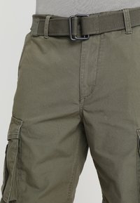 Pantaloni cargo in tessuto verde oliva con una texture liscia, dotati di una cintura ampia, tasche laterali e una vestibilità slim.