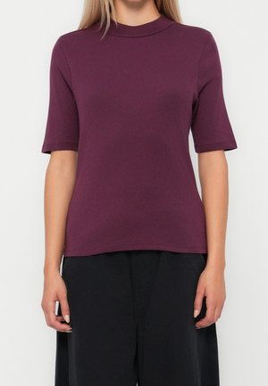 Kobieta ubrana w dopasowany, krótki rękaw bluzki w kolorze śliwkowym z wysokim okrągłym dekoltem oraz luźne czarne spodnie.