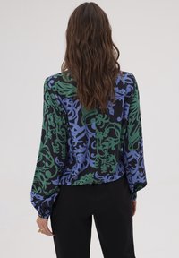 Blouse à manches longues avec un motif floral noir, vert et bleu, décolleté en V et poignets froncés, accompagnée d'un pantalon noir. Matière douce, coupe décontractée.