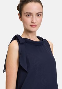 Námornícka bezrukávová blúzka s prehnutým, zaobleným výstrihom a látkovými mašľami na ramenách. Vyrobená z ľahkého, hladkého materiálu s jemným leskom.