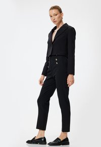 Blazer nero cropped con un unico bottone, abbinato a pantaloni neri a vita alta con bottoni dorati e tasche laterali. Mocassini neri.