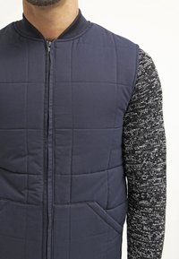 Marinblå kviltad väst med dragkedja fram, rund krage och två sidofickor; matchad med en svart och vit texturerad långärmad skjorta.