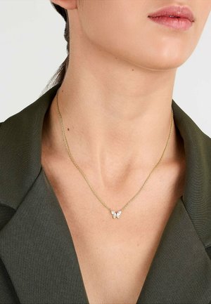 Nahaufnahme einer Person, die eine goldene Halskette mit einem kleinen schmetterlingsförmigen Anhänger trägt, gekleidet in einen dunkelgrünen Blazer.