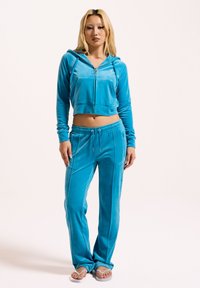 Abgeschnittene blaue Velours-Trainingsjacke mit Kapuze, kombiniert mit passenden Sweatpants mit Kordelzug. Geschmeidige Textur mit dezenten Nahtdetails.