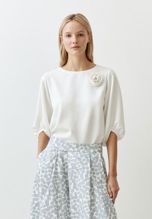 Weißes Bluse mit Rundhalsausschnitt und Puffärmeln, akzentuiert mit einem strukturierten Blumenmotiv; kombiniert mit einem hellblauen gemusterten Rock.
