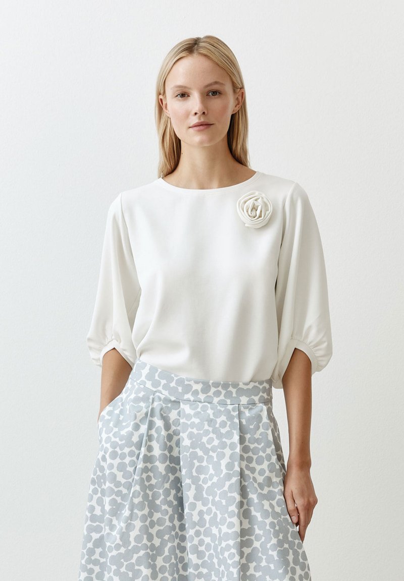 Witte blouse met een ronde hals en pofmouwen, verfraaid met een getextureerd bloemdetail; gecombineerd met een lichtblauwe gestreepte rok.