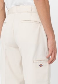 Pantalones color beige claro con una textura suave, que cuentan con dos bolsillos traseros y un bolsillo lateral tipo cargo con un parche de logotipo de marca colorido.