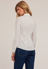 Blusa blanca con un diseño ajustado, mangas largas y cuello puntiagudo. Confeccionada en tejido suave con acentos de botones en los puños. Llevada con jeans de mezclilla azules.