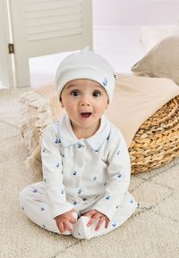 Vit heldräkt med ett blått båt-mönster, knäppning med knappar och en matchande vit hatt. Mjuk tyg, långa ärmar och fotad design.
