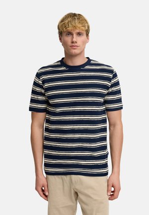 Junger Mann mit blondem Haar, der ein marineblau-weiß gestreiftes Kurzarmhemd und beige Hosen trägt, steht vor einem schlichten Hintergrund.