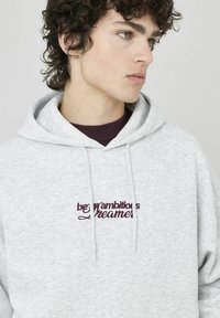 Sweat à capuche gris fabriqué en tissu doux et texturé. Il comporte une poche avant et un texte brodé bordeaux qui dit "soyez des rêveurs ambitieux".