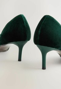 Pantofi cu toc înalt din catifea verde, cu vârfuri ascuțite, având un design elegant și tocuri subțiri. Textura este moale și luxoasă.