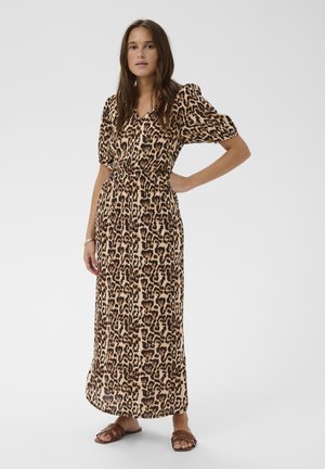 Žena stojaca s rukou na bokoch, oblečená v šatách s leopardím vzorom dĺžky po členky, s nafúknutými krátkymi rukávmi a hnedými sandálmi.