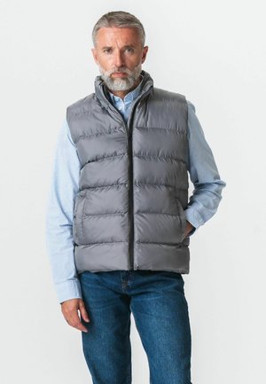 Mężczyzna w średnim wieku z siwą brodą, ubrany w szarą pikowaną kamizelkę na niebieskiej koszuli w paski i niebieskie dżinsy, stojący na jednolitym tle.