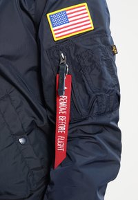 Mørkeblå bomberjakke med et amerikansk flag med gul kant, rød "FJERN FØR FLYVNING" mærkat og struktureret nylonstof.