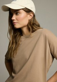 Beżowy, oversize T-shirt z okrągłym dekoltem, w połączeniu z kremową czapką baseballową z haftowanym napisem. Długie, falujące włosy częściowo widoczne.