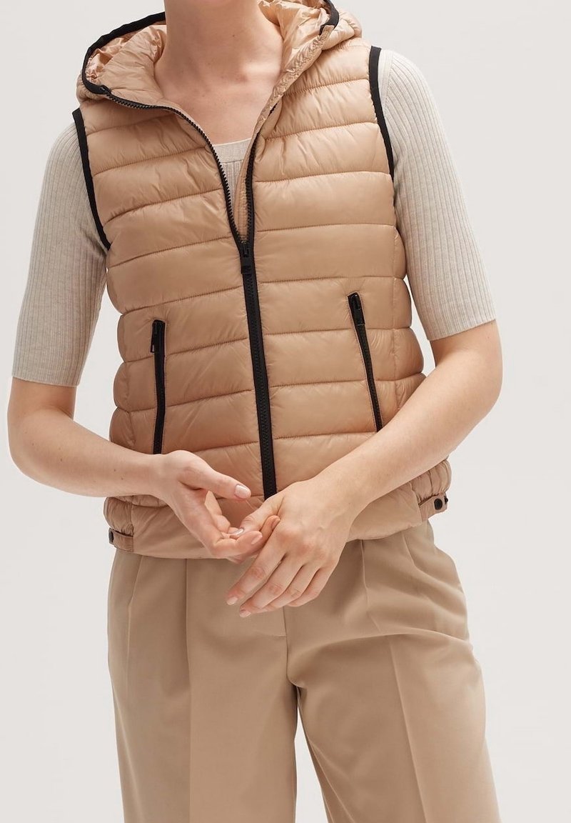 Beige pufferväst med huva, med vertikal sömnad, svarta dragkedjor och detaljer, i kombination med en beige åtsittande topp och byxor.