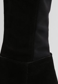 Gros plan sur la tige de la botte en suède noir et tissu, montrant les coutures détaillées et le contraste de textures entre les matériaux.