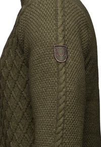 Olivgrön stickad tröja med en texturerad mönster, kabeldetaljer och ett litet logopatch på axeln.
